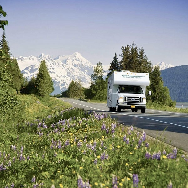 RV traveling an Alaska highway past wildflowers and snow-capped mountains
