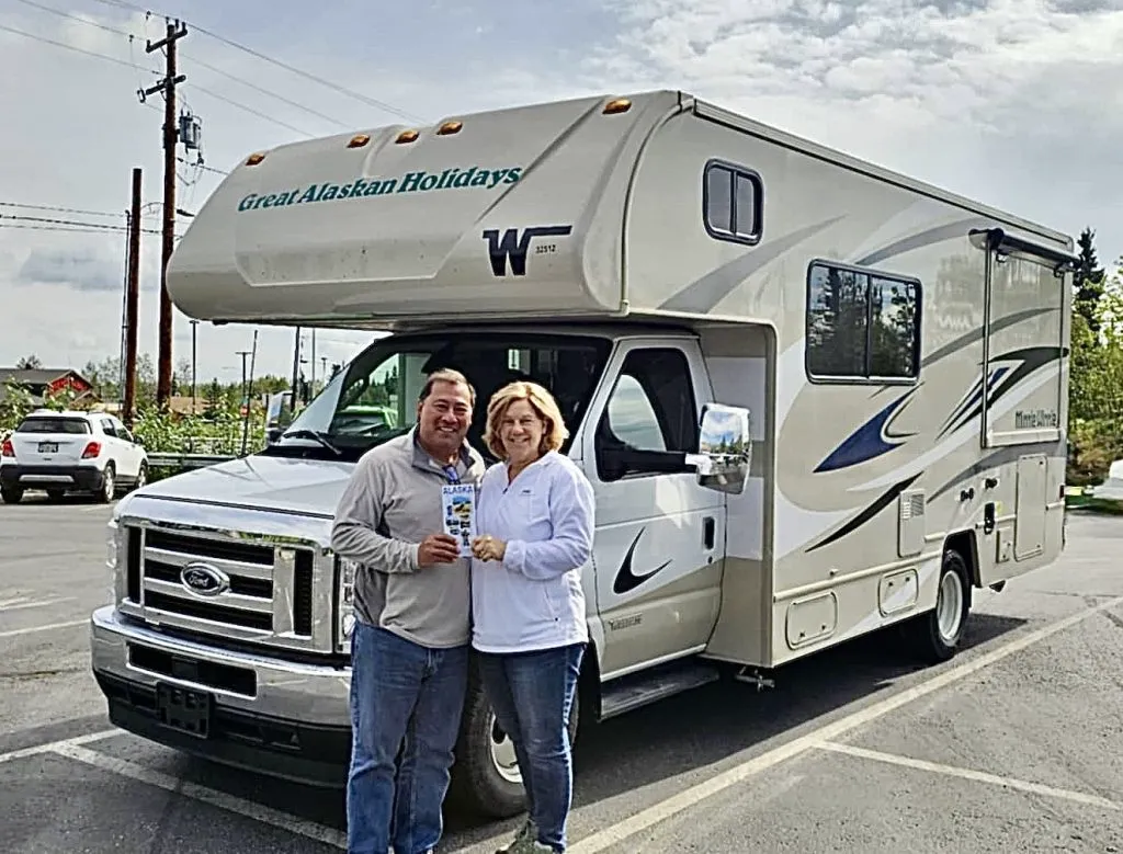 A smiling couple holding the printed Alaska campground guide in front of a Great Alaskan Holidays motorhome
